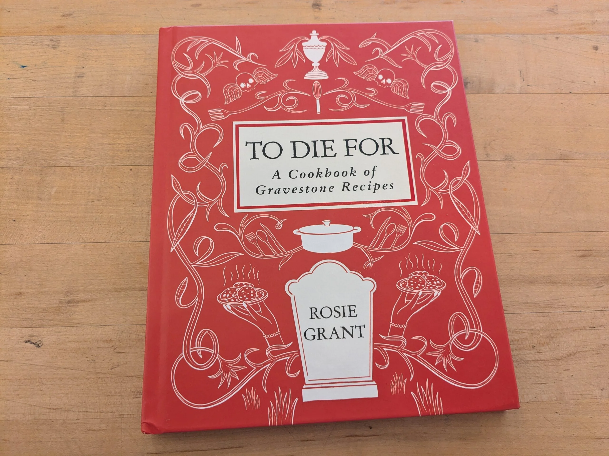 A red hardcover book titled “To Die For: A Cookbook of Gravestone Recipes” by Rosie Grant is on a wooden surface. The cover features white illustrations of gravestones and food.