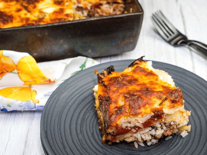 A slice of baked casserole with rice, vegetables, and a browned cheese topping is served on a black plate, with the remaining dish visible in the background.