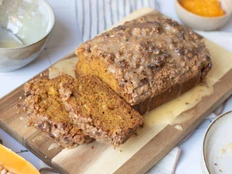 A loaf of pumpkin bread with a crumb topping and glaze sits on a wooden board, with two slices cut. Bowls are visible in the background.