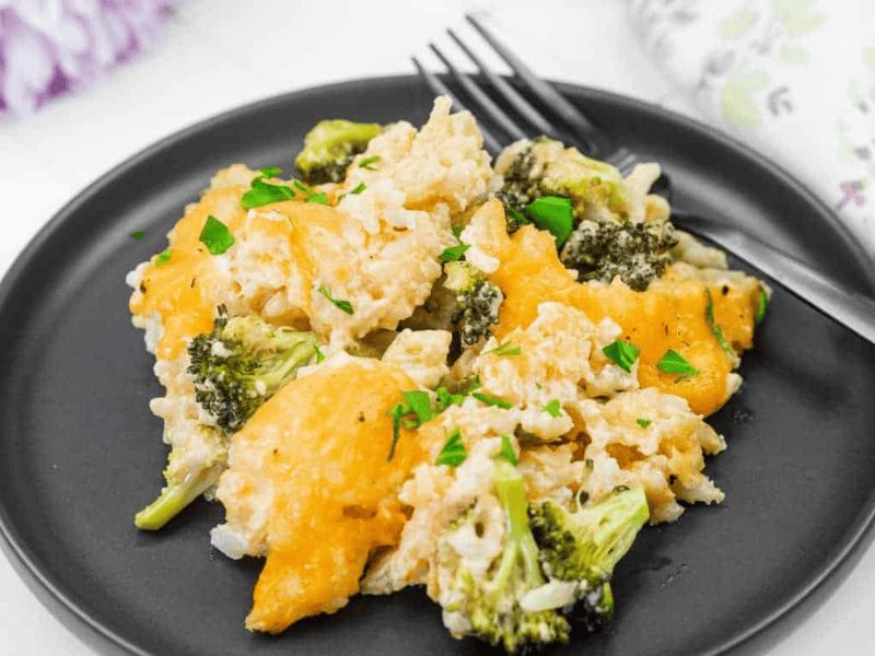 A serving of broccoli and cheese casserole with rice on a black plate, garnished with parsley, and a fork placed beside the food.