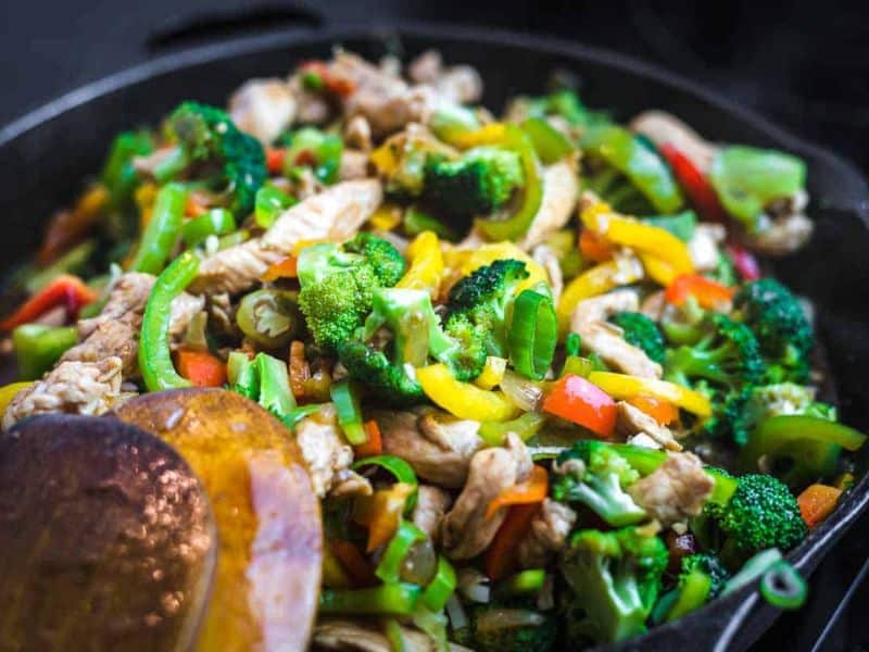 A close-up of a stir-fry dish with mixed vegetables, including broccoli, bell peppers, and green onions, and pieces of cooked chicken in a pan with a wooden spoon.