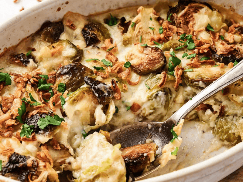 Casserole dish with creamy Brussels sprouts, topped with crispy onions and chopped herbs, with a serving spoon in the dish.