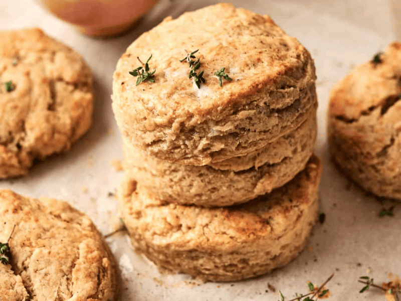 Two golden brown biscuits are stacked on parchment paper, with a sprig of fresh thyme on top. Other biscuits are visible nearby.