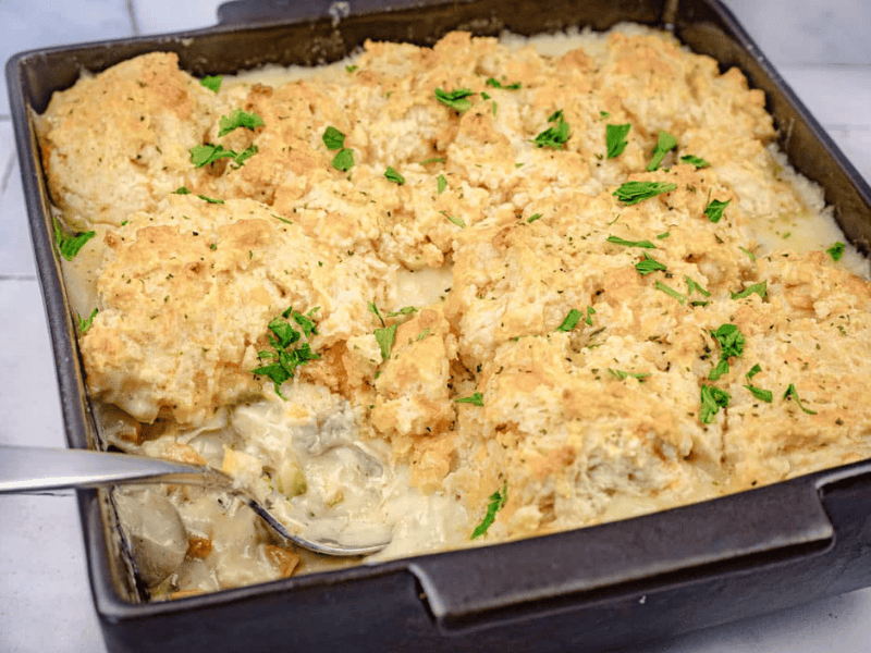 A baking dish of chicken and biscuit casserole topped with golden biscuits and garnished with chopped parsley, with a serving spooned out.