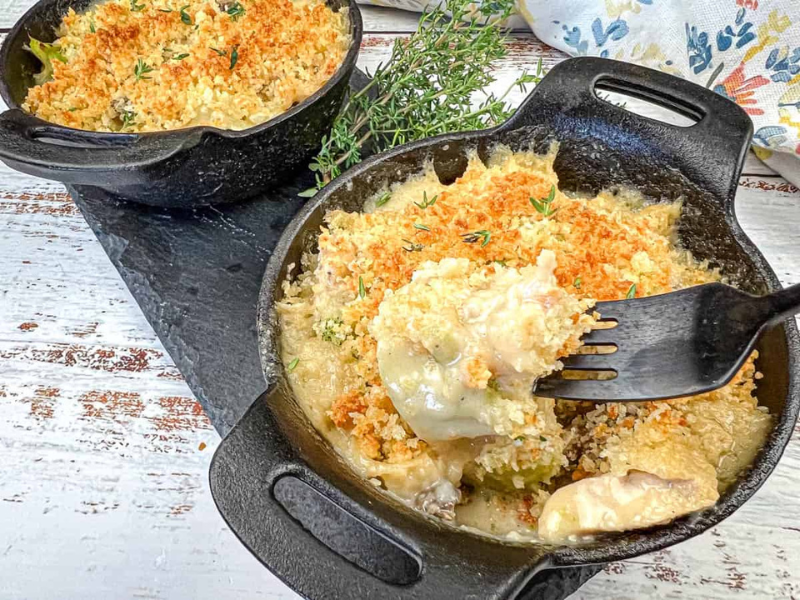 Two small cast iron skillets filled with creamy baked casserole topped with golden breadcrumbs, garnished with fresh thyme, on a rustic white surface.