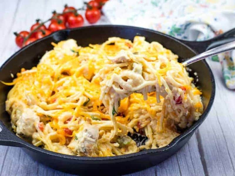 A fork lifts a portion of cheesy pasta from a black skillet, with cherry tomatoes and a floral napkin in the background.