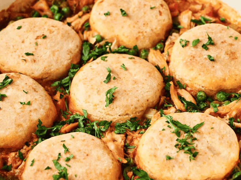 Close-up of a casserole topped with six round biscuits, shredded chicken, peas, and chopped parsley.