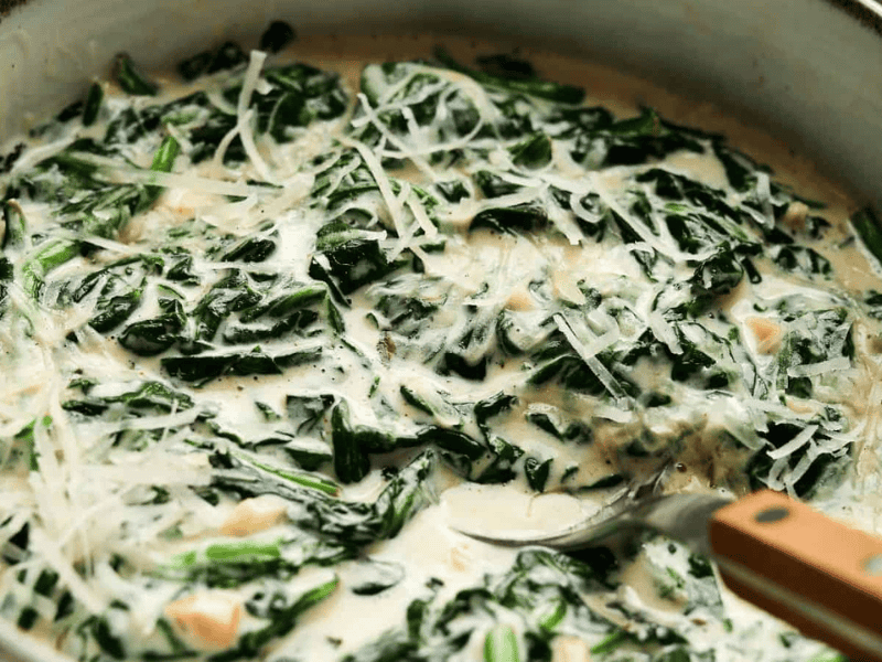 A close-up of creamy spinach with shredded cheese in a pan, being stirred with a wooden-handled spoon.
