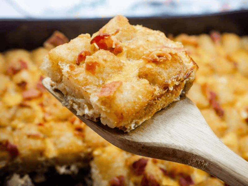 A wooden spatula holds a square serving of baked casserole topped with tater tots and bits of bacon, with the remainder of the casserole in the background.