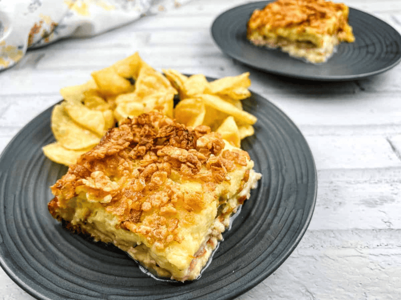 A slice of casserole topped with crispy cereal served with potato chips on a black plate, with another similar plate in the background.