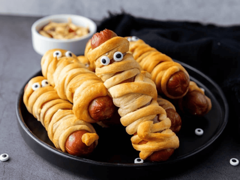 A plate of sausage mummies made from hot dogs wrapped in dough, baked golden brown, with candy eyes, and a small bowl of dipping sauce in the background.
