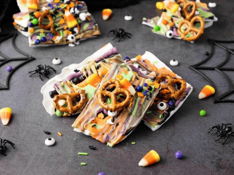 Colorful Halloween bark topped with pretzels, candy eyes, sprinkles, and candy corn, arranged on a dark surface with plastic spiders and web decorations.