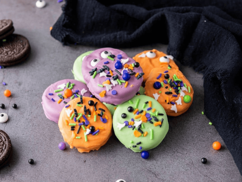 Colorful Halloween-themed cookies with purple, orange, and green icing, decorated with sprinkles and candy eyes, arranged on a gray surface near a black cloth.