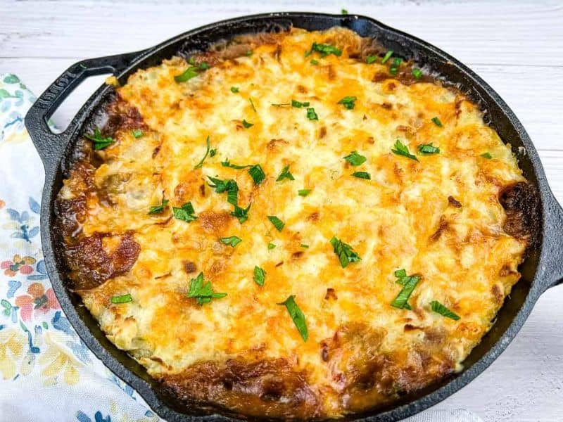 A baked casserole topped with melted, golden-brown cheese and garnished with chopped parsley in a cast iron skillet.
