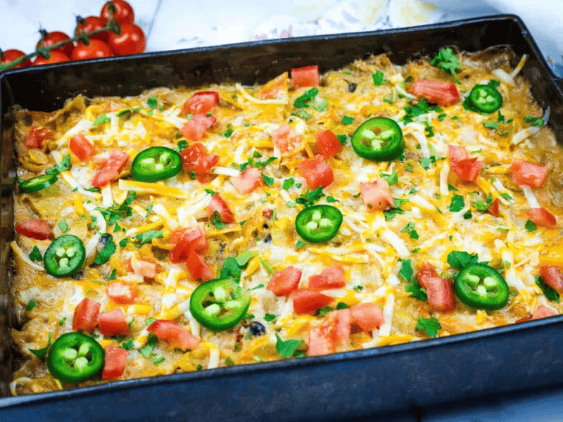 A baked casserole topped with melted cheese, sliced jalapeños, chopped tomatoes, and fresh herbs in a rectangular pan.