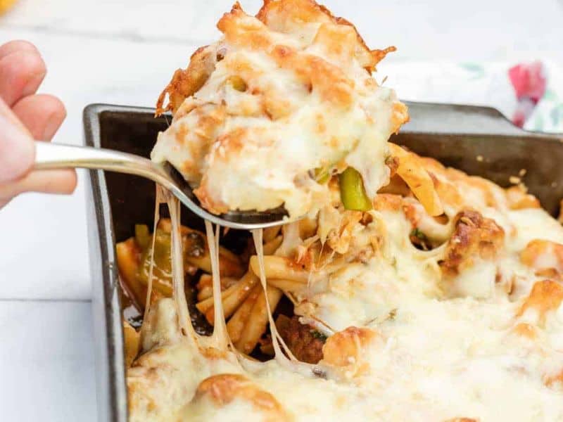 A spoon lifts a serving of baked casserole topped with melted cheese from a rectangular pan, showing pasta and vegetables underneath.