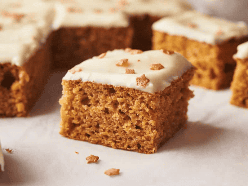A piece of iced ginger cake with white frosting and small star-shaped sprinkles, placed on parchment paper with more pieces in the background.