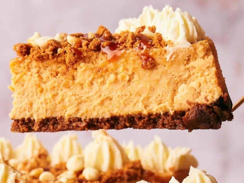 A slice of creamy cheesecake with a brown crumb crust, whipped cream topping, caramel drizzle, and crumbled cookie pieces is being lifted from a whole cheesecake.