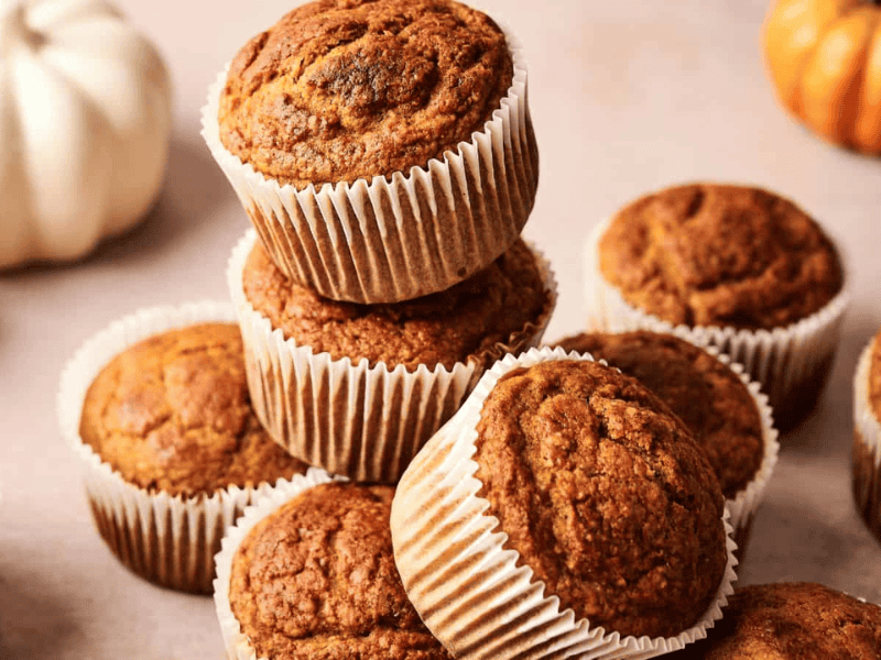 A group of pumpkin muffins in white paper liners are stacked and arranged on a light-colored surface.