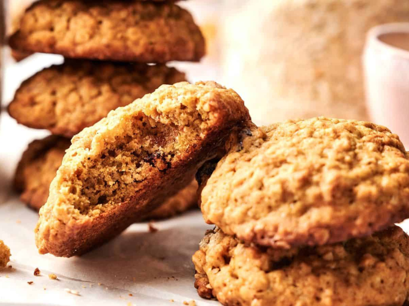 Close-up of several oatmeal cookies, with one cookie broken in half to show the texture inside. Other cookies are stacked and placed on a white surface.
