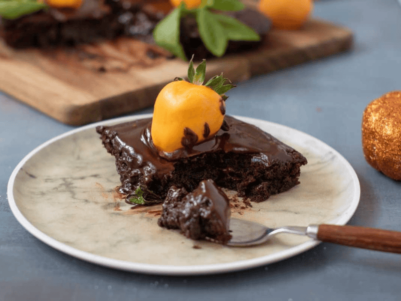 A slice of chocolate cake topped with chocolate sauce and a yellow fruit is served on a plate with a fork; more cake is visible in the background.