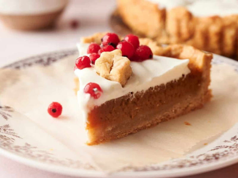 A slice of pie with a creamy topping, biscuit pieces, and red berries is served on a patterned plate.