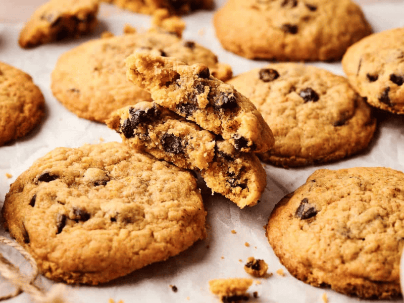 A close-up of several chocolate chip cookies on parchment paper, with one cookie broken in half to show the inside.