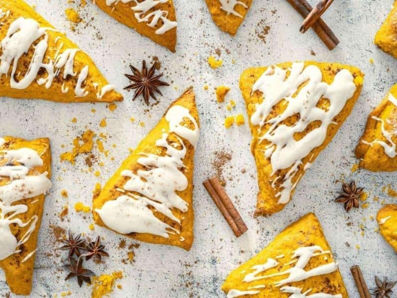 Pumpkin scones with white icing are arranged on a light surface, surrounded by cinnamon sticks and star anise.