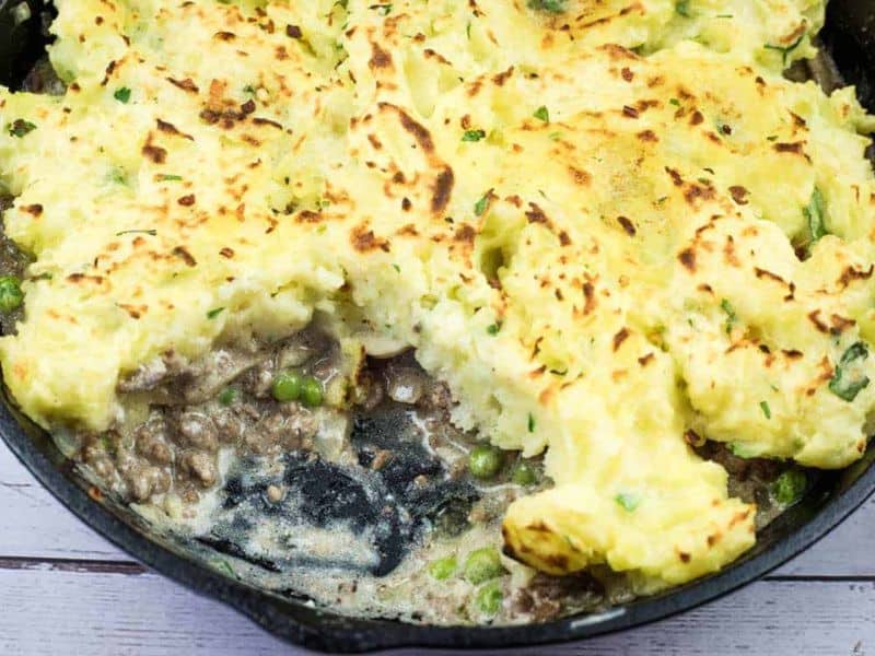A partially served skillet of shepherd's pie with browned mashed potato topping, ground meat, peas, and sauce visible inside.