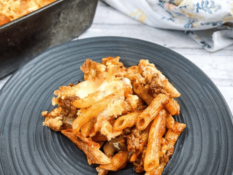 A serving of baked ziti with ground meat, tomato sauce, and melted cheese on a black plate, with a casserole dish and napkin in the background.