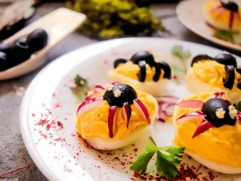 Deviled eggs topped with black olive and red vegetable pieces arranged to look like spiders, garnished with parsley, on a white plate.