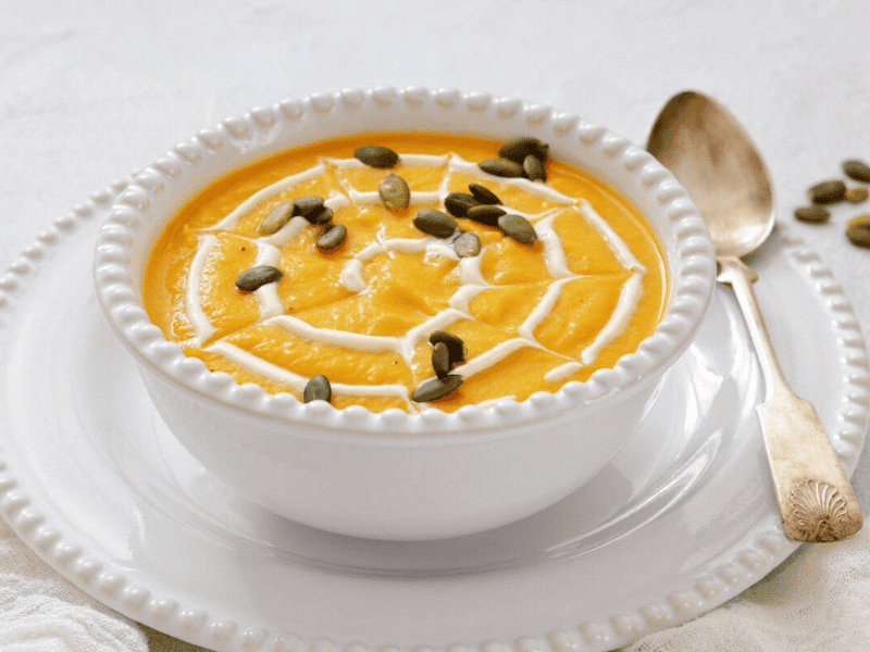 A white bowl of creamy pumpkin soup topped with a swirl of cream and pumpkin seeds, placed on a white plate with a spoon beside it.