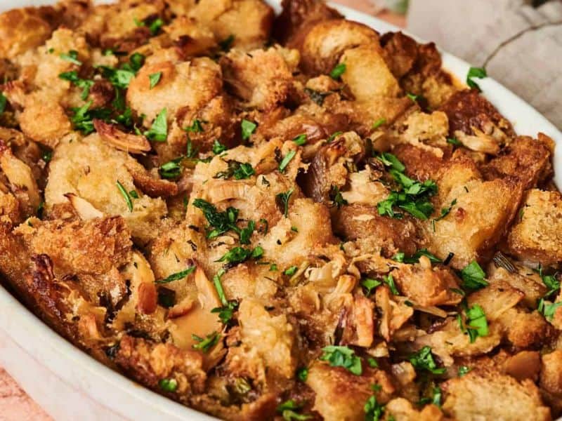 Close-up of baked bread stuffing in a white dish, topped with chopped parsley and visible chunks of bread and herbs.