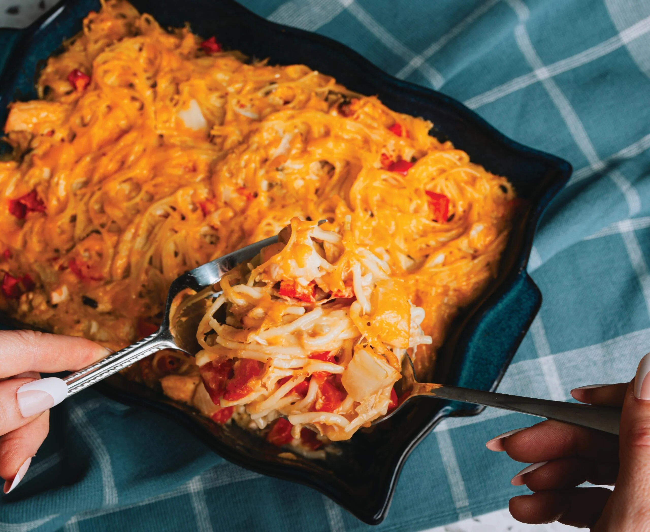 A person serves a cheesy pasta bake with visible chunks of chicken and diced tomatoes from a baking dish using a large spoon.