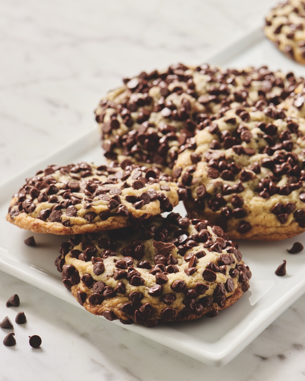 A plate of large chocolate chip cookies is shown on a white marble surface, with extra chocolate chips scattered nearby.