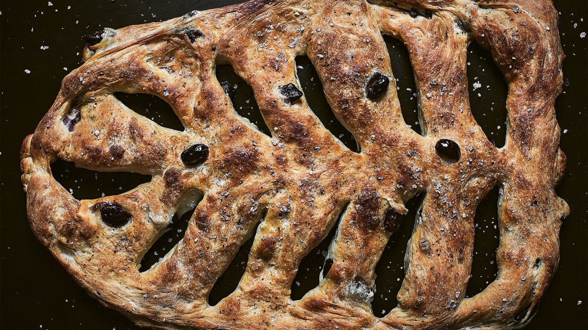 A flatbread topped with salt and black olives, with decorative openings cut into the dough on the diagonal. It's on a black background. 