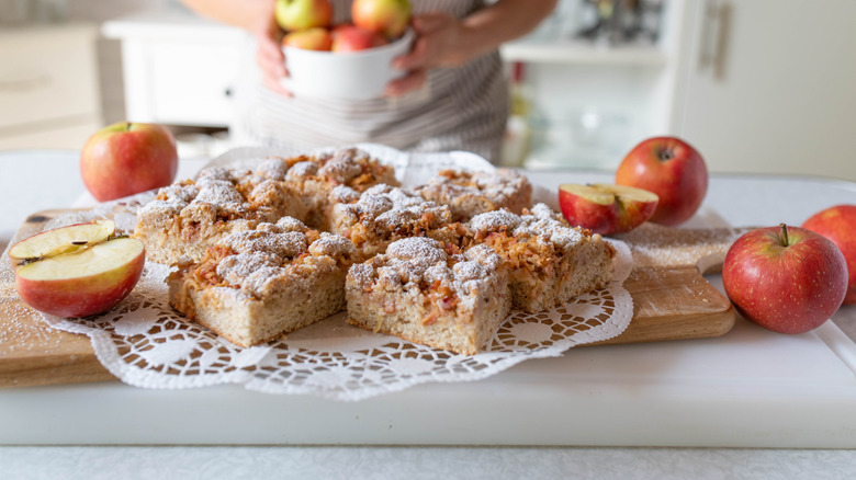 Pieces of apple sheet cake on a cutting board beside sliced apples.