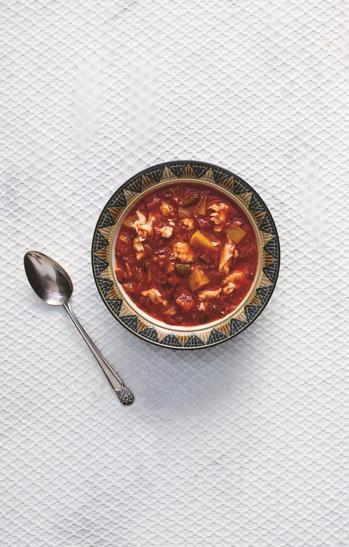 3 Comforting Stews from Michael W. Twitty's New Cookbook - 2 A patterned bowl filled with chunky tomato-based soup sits next to a spoon on a white textured surface.