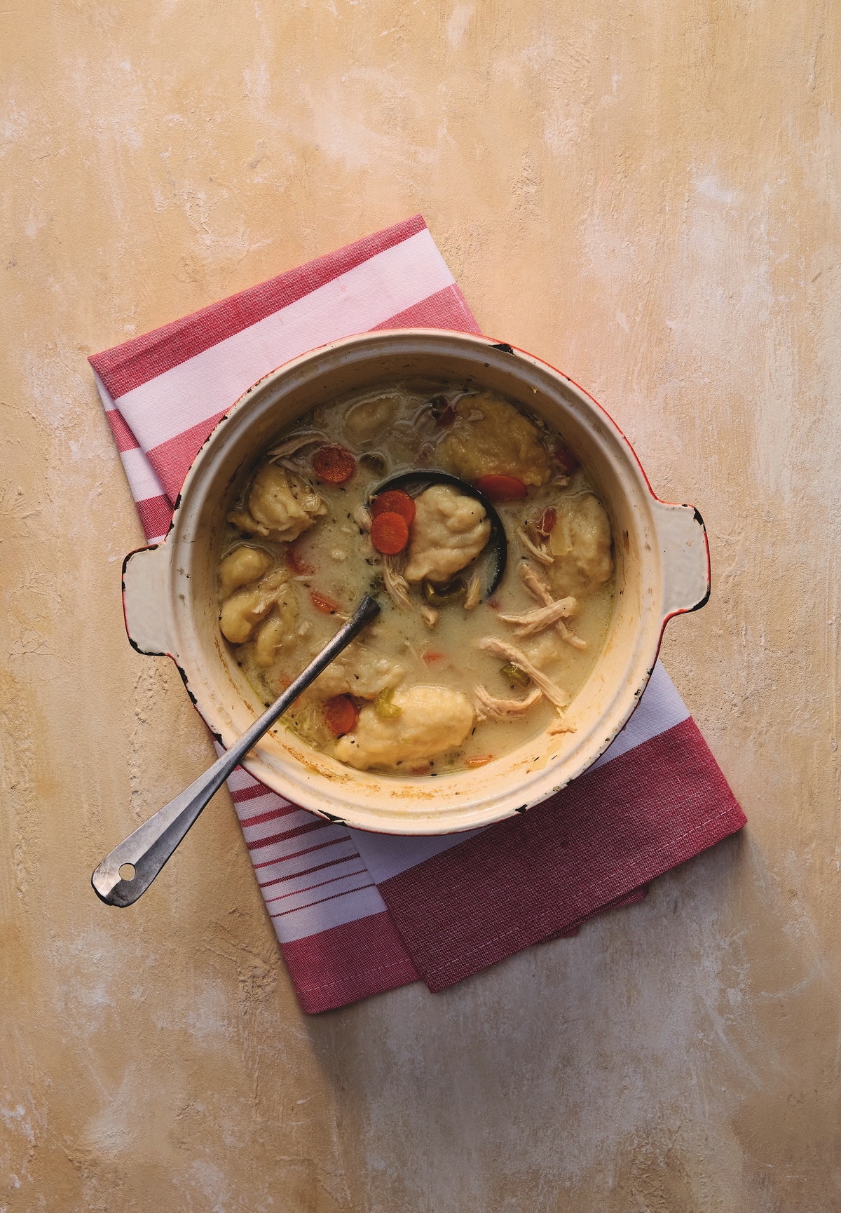 3 Comforting Stews from Michael W. Twitty's New Cookbook - 1 A pot of chicken and dumplings with carrots and a ladle, set on a red and white striped towel on a beige surface.