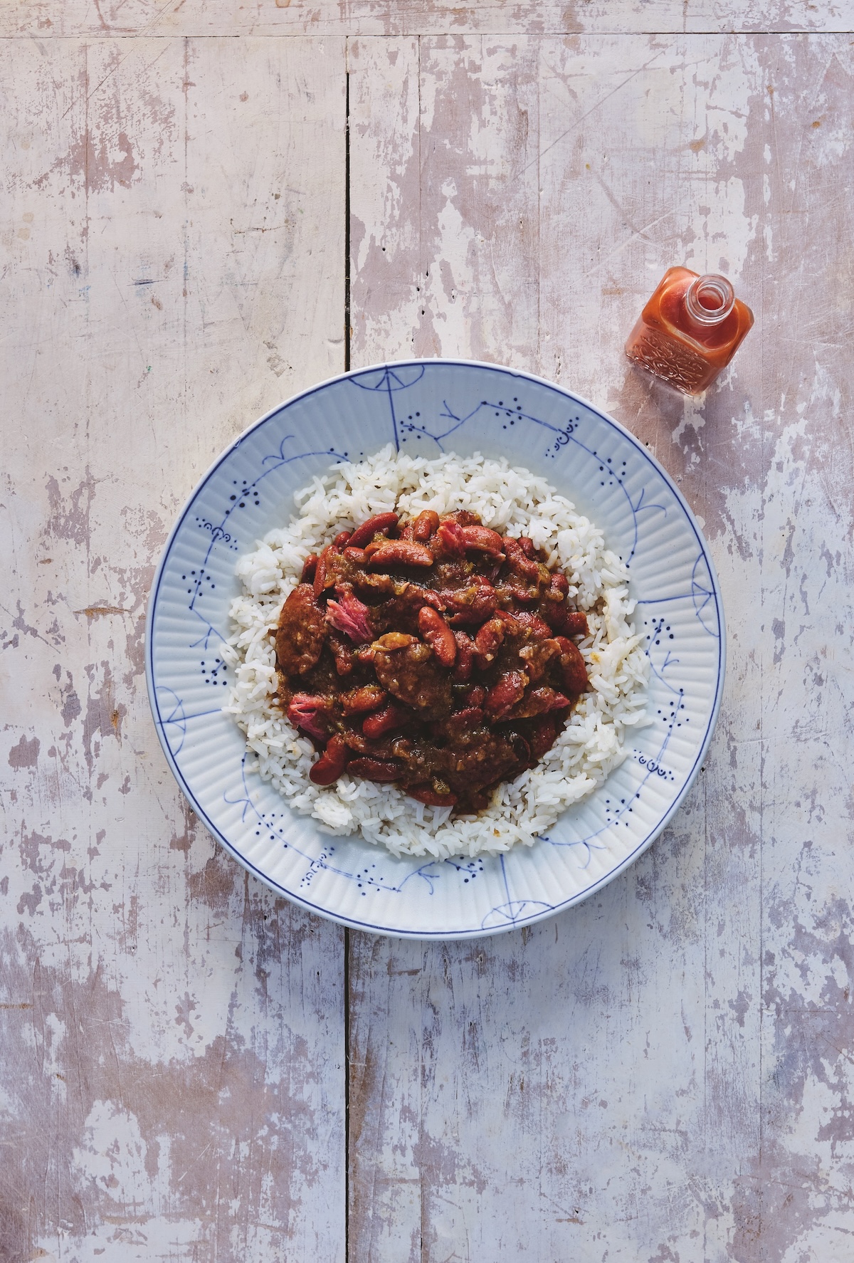 3 Comforting Stews from Michael W. Twitty's New Cookbook - 3 A plate of white rice topped with a stew of red beans and meat, placed on a rustic wooden surface, with a small bottle of hot sauce beside it.